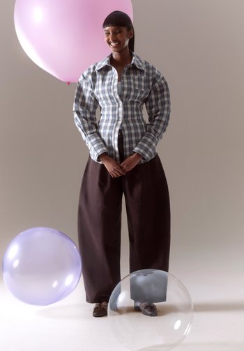 Femme souriante, portant une chemise à carreaux et un pantalon large et sombre, debout parmi de grands ballons aux couleurs pastel sur un fond neutre.