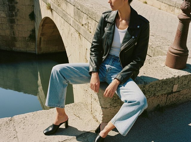 Jeune personne portant une veste en cuir noire, un haut blanc et un jean bleu clair, assise sur un rebord en pierre près d'un canal avec un pont en pierre voûté.