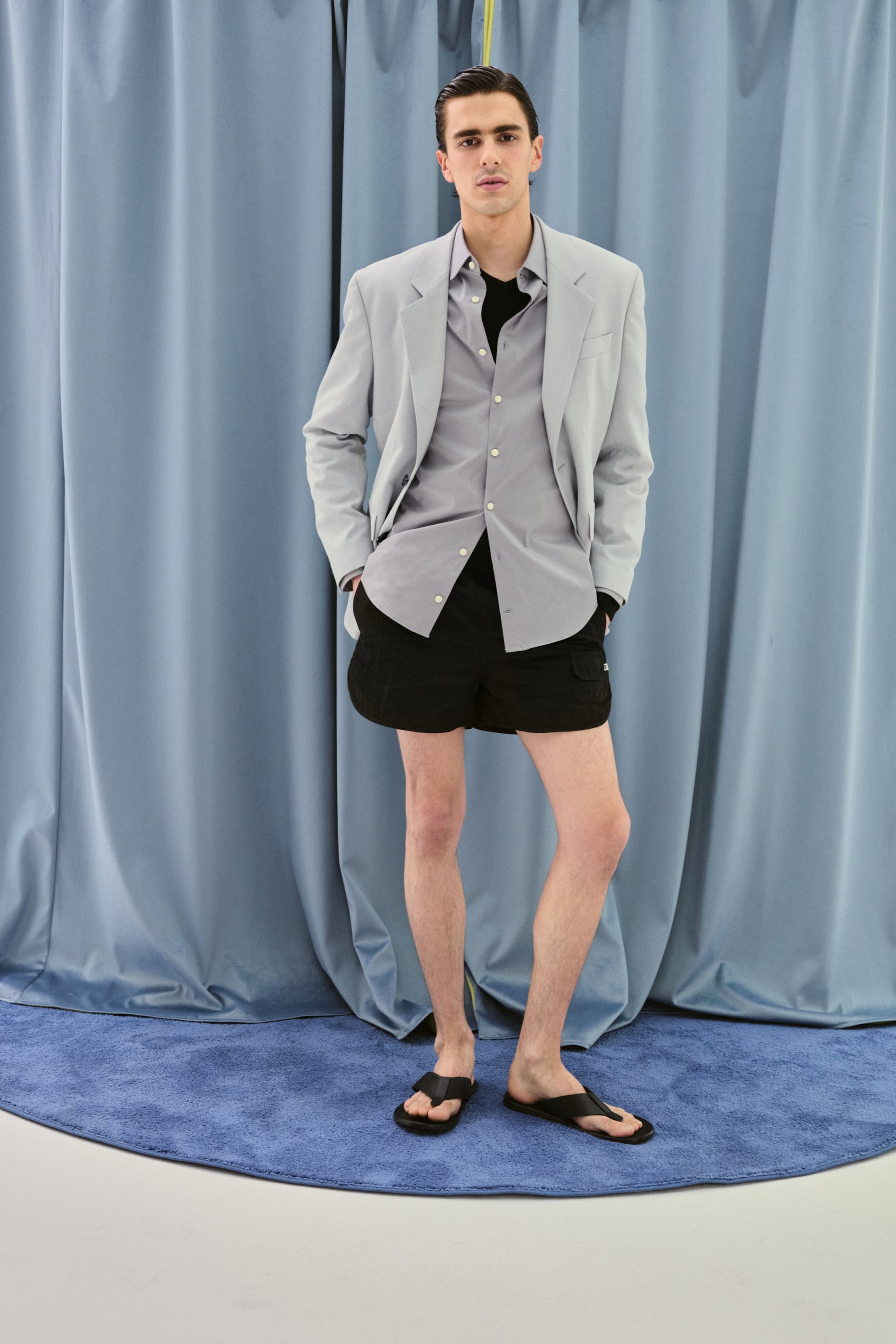 Un giovane uomo sta in piedi con sicurezza su un tappeto blu circolare, indossando un blazer grigio chiaro, una camicia scura, pantaloni corti neri e infradito.