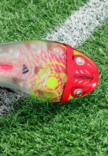 Semelle claire d'une chaussure de football rouge et jaune reposant sur une pelouse artificielle verte avec une ligne blanche de terrain.