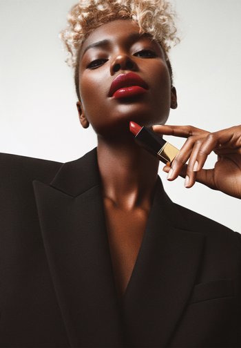 Rouge à lèvres avec un étui métallique noir et doré. Modèle portant un blazer noir oversize, avec des cheveux blonds courts et bouclés et des lèvres brillantes.
