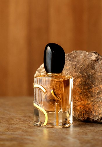 Flacon de parfum transparent avec bouchon noir et accents dorés à côté d'un rocher texturé sur une surface et un fond marron.