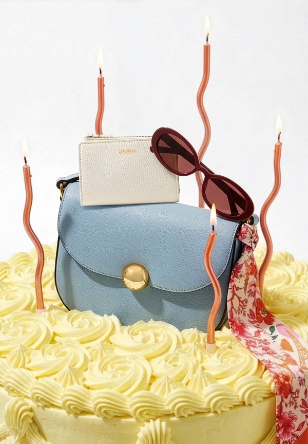 Light blue leather handbag with floral scarf, white wallet, and brown sunglasses on a cream frosted cake with seven lit wavy candles.
