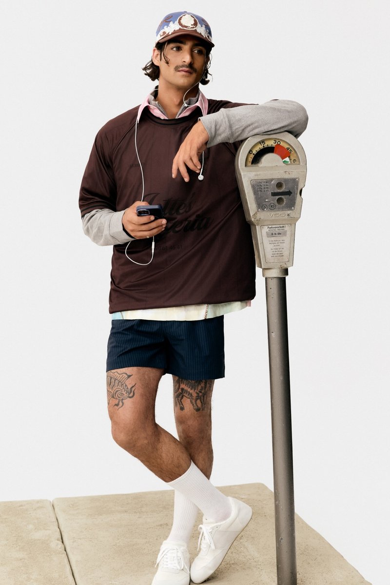 Ein junger Mann mit einem Schnurrbart lehnt an einem Parkscheinautomaten. Er trägt ein dunkles Hemd, gestreifte Shorts und weiße Turnschuhe und hält ein Handy in der Hand.