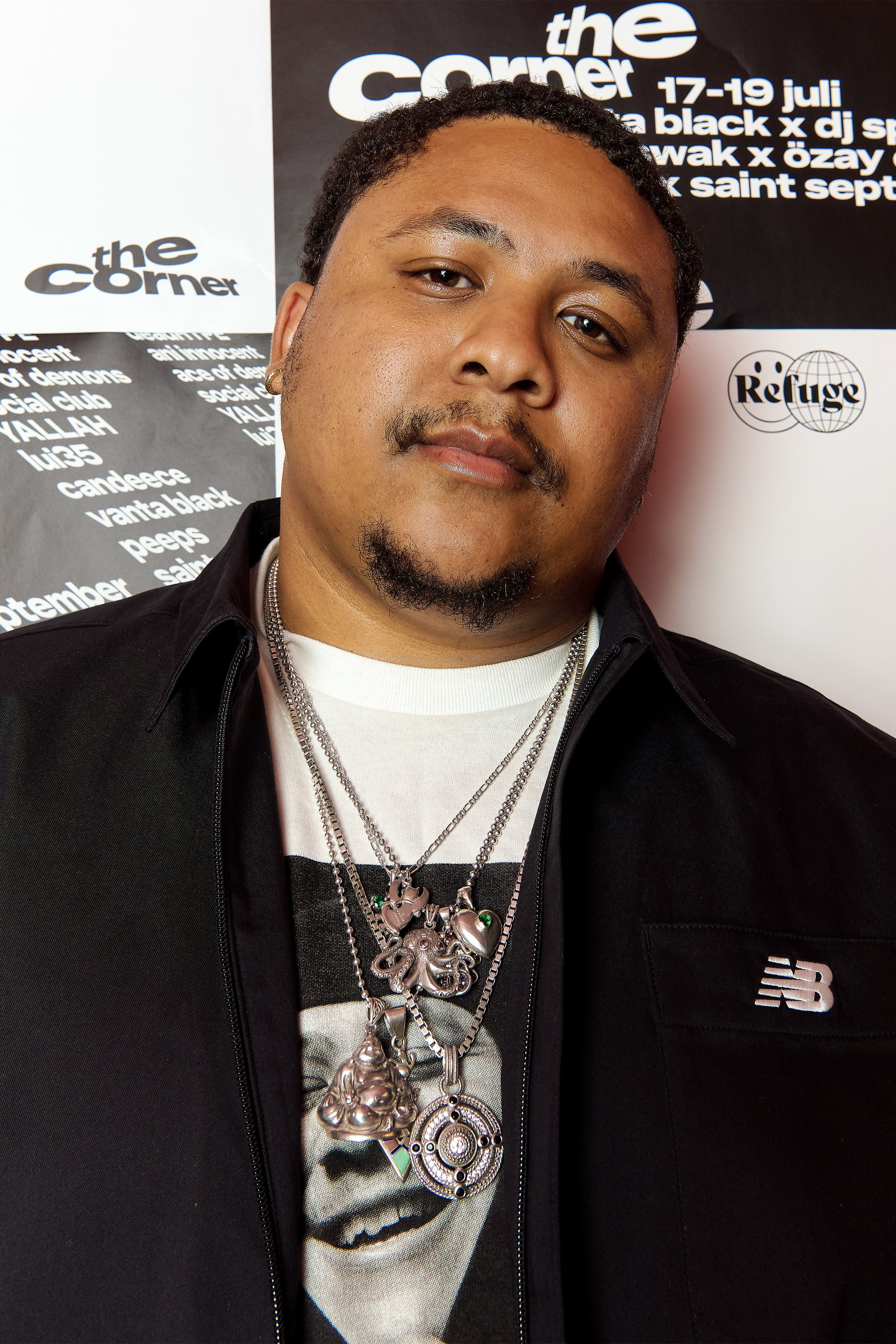 Mand med kort krøllet hår og skæg, iført sort jakke og flere sølvringe med forskellige vedhæng mod en promoveringsbaggrund.