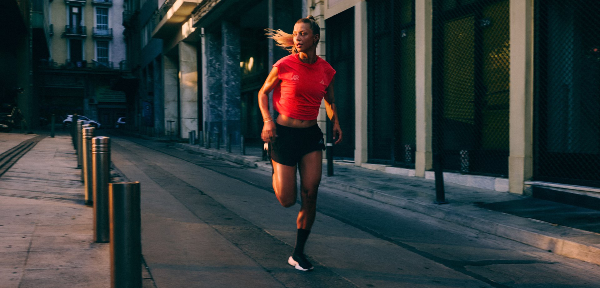 Runner in red shirt and black shorts jogging on an urban street in dim light with buildings and bollards along the sidewalk.