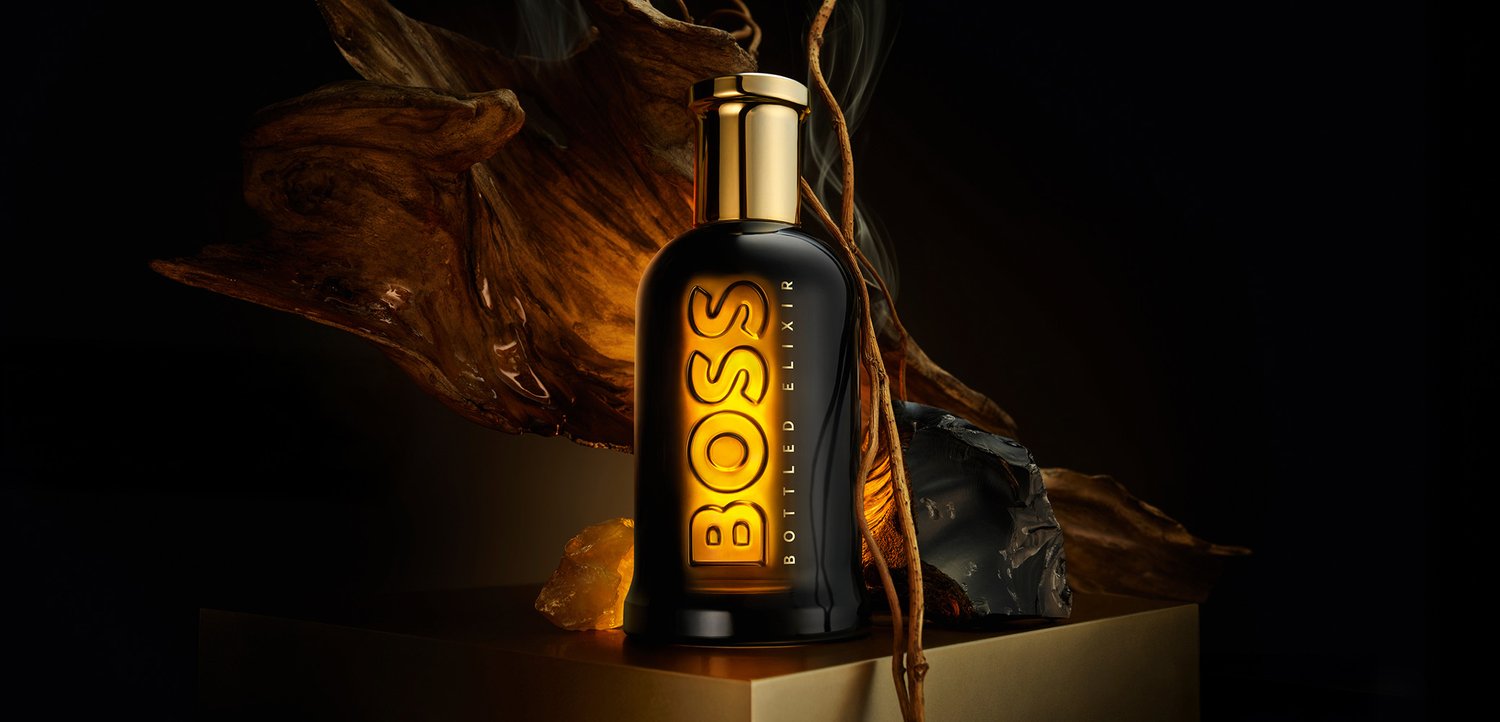 Flacon de parfum BOSS Bottled Elixir noir et or sur un socle avec un éclairage dramatique et des éléments naturels en bois et ambre.