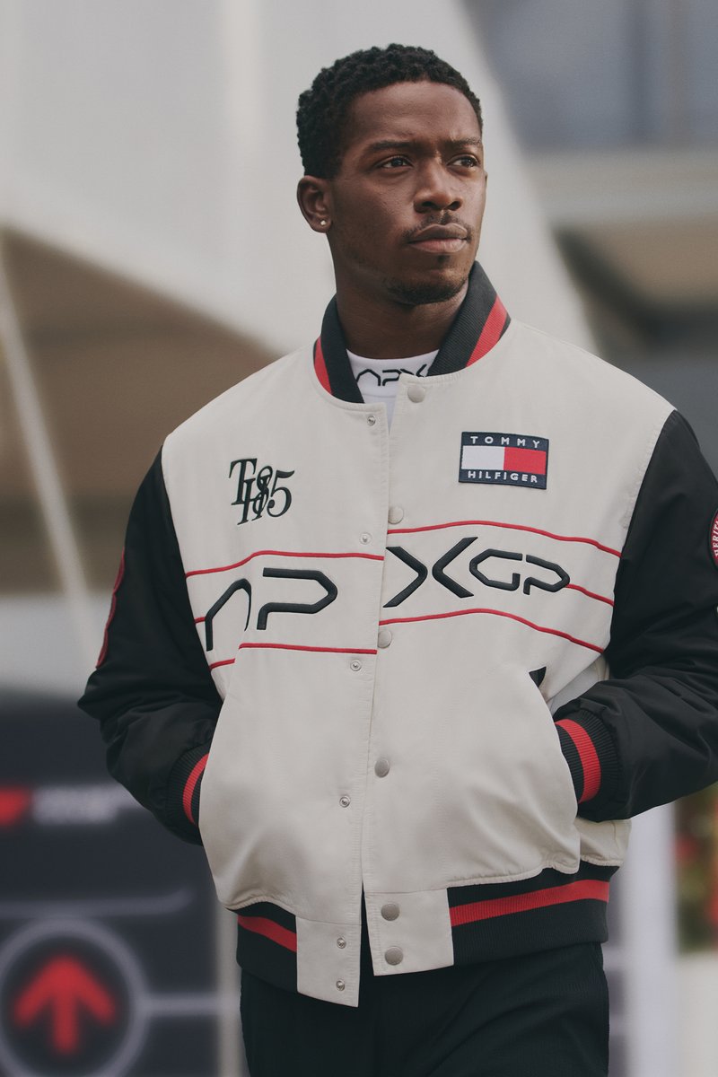 A man with short hair wears a stylish white and black bomber jacket with red accents, standing confidently against a modern backdrop.