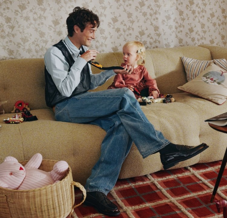 Beige tygsoffa med texturerad yta. En man i svart väst och blå skjorta interagerar med en flicka i rosa topp, omgiven av leksaker.