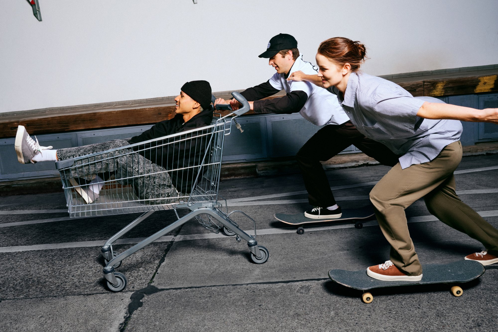 Three young people skateboarding in a parking area; one sits in a shopping cart while the others push and balance on skateboards.