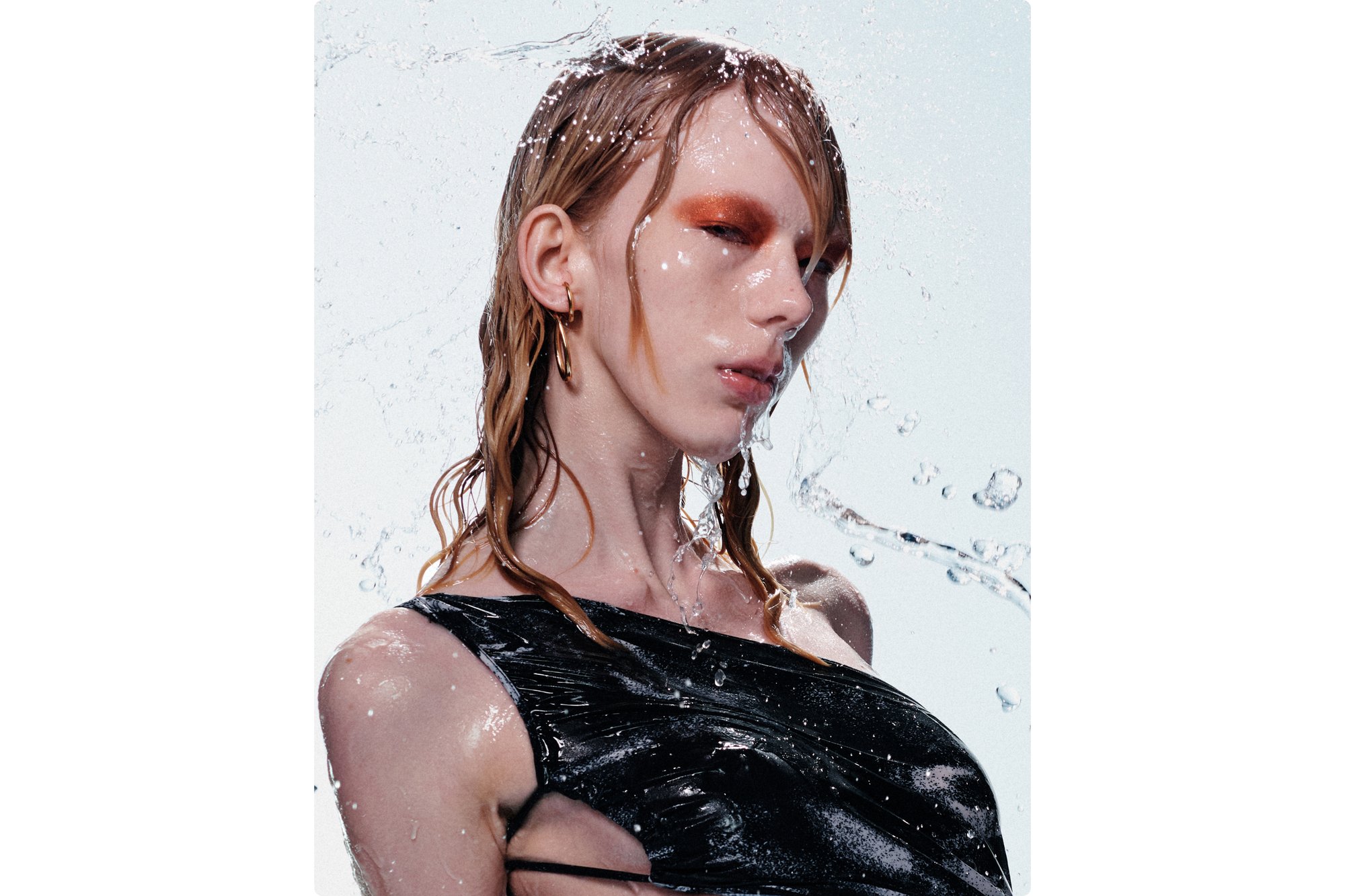 Een model met nat haar en gedurfde oranje oogschaduw kijkt intens terwijl water om haar heen spat, gekleed in een zwart, aansluitend kledingstuk.