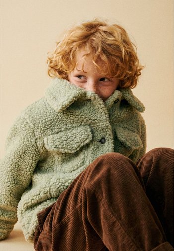 Enfant roux avec des tâches de rousseur portant une veste verte duveteuse et un pantalon en velours côtelé marron, assis et regardant sur le côté avec une expression timide.