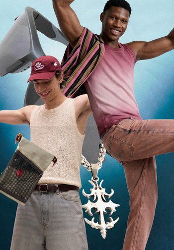 Zwei junge Männer lächeln und posieren; einer trägt eine rote Kappe und ein Netz-Tanktop mit einer Umhängetasche, der andere trägt einen gestreiften Schal, ein pinkes Tanktop und eine Hose.