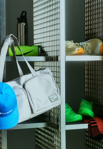 Biała torba sportowa Nike zawieszona na metalowej półce z niebieską czapką Champion, zieloną butelką na wodę, beżowo-zielonymi butami sportowymi oraz pomarańczowo-paskowanymi skarpetkami.