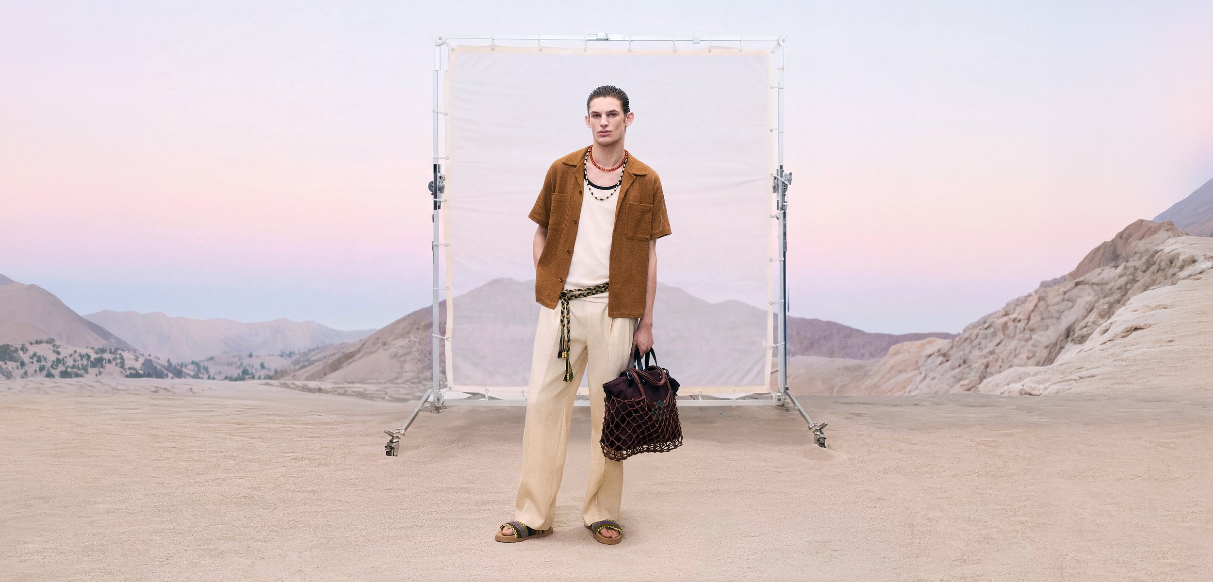 Joven de pie en terreno desértico con ropa casual beige y marrón, sosteniendo una gran bolsa tejida, con montañas y un fondo blanco detrás de él.