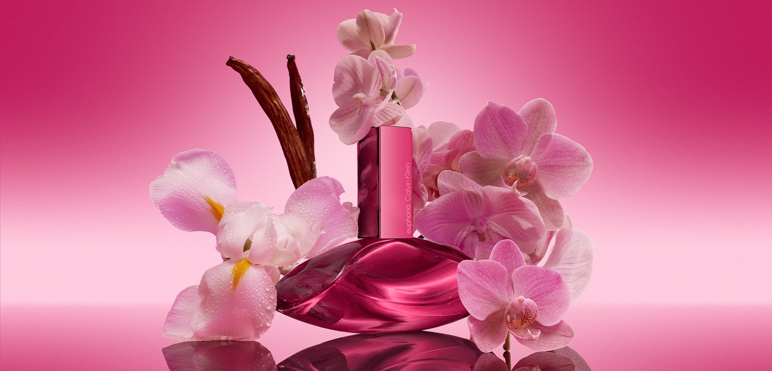 Pink parfume flaske omgivet af pink orkideer og vaniljestænger på en reflekterende overflade med en pink gradient baggrund.
