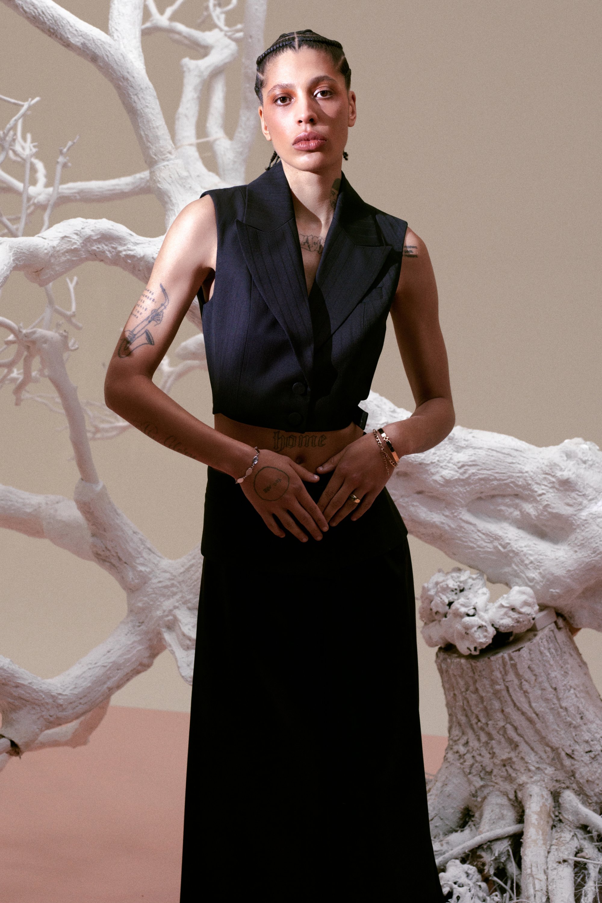 Persona con trenzas posa en un atuendo negro sin mangas, mostrando tatuajes, frente a un fondo marrón claro y ramas de árbol abstractas en blanco.