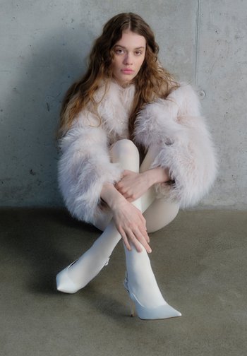 Un model șezând pe o suprafață netedă, purtând o haină pufoasă roz deschis, ciorapi albi transparenți și pantofi cu toc ascuțiți, de culoare pală.