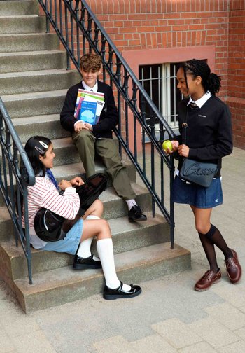 Trois élèves en uniforme scolaire interagissent sur des escaliers extérieurs ; l'un tient des livres, un autre joue de la guitare et un autre tient une pomme verte.