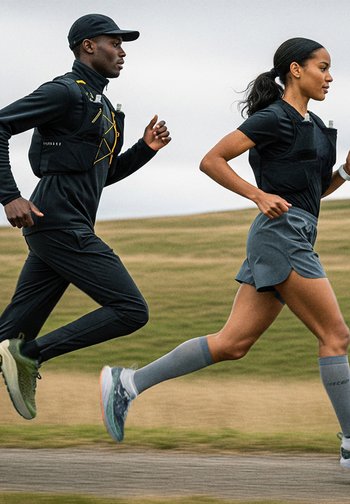 Due corridori in abbigliamento sportivo nero, che indossano gilet idratanti. L'uomo ha pantaloni neri e sneakers olive; la donna ha una gonne grigia e calzini.