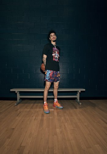 Un jeune homme en tenue de basketball colorée se tient dans une salle de gym, tenant un ballon de basket, avec un banc à proximité et un mur sombre en arrière-plan.