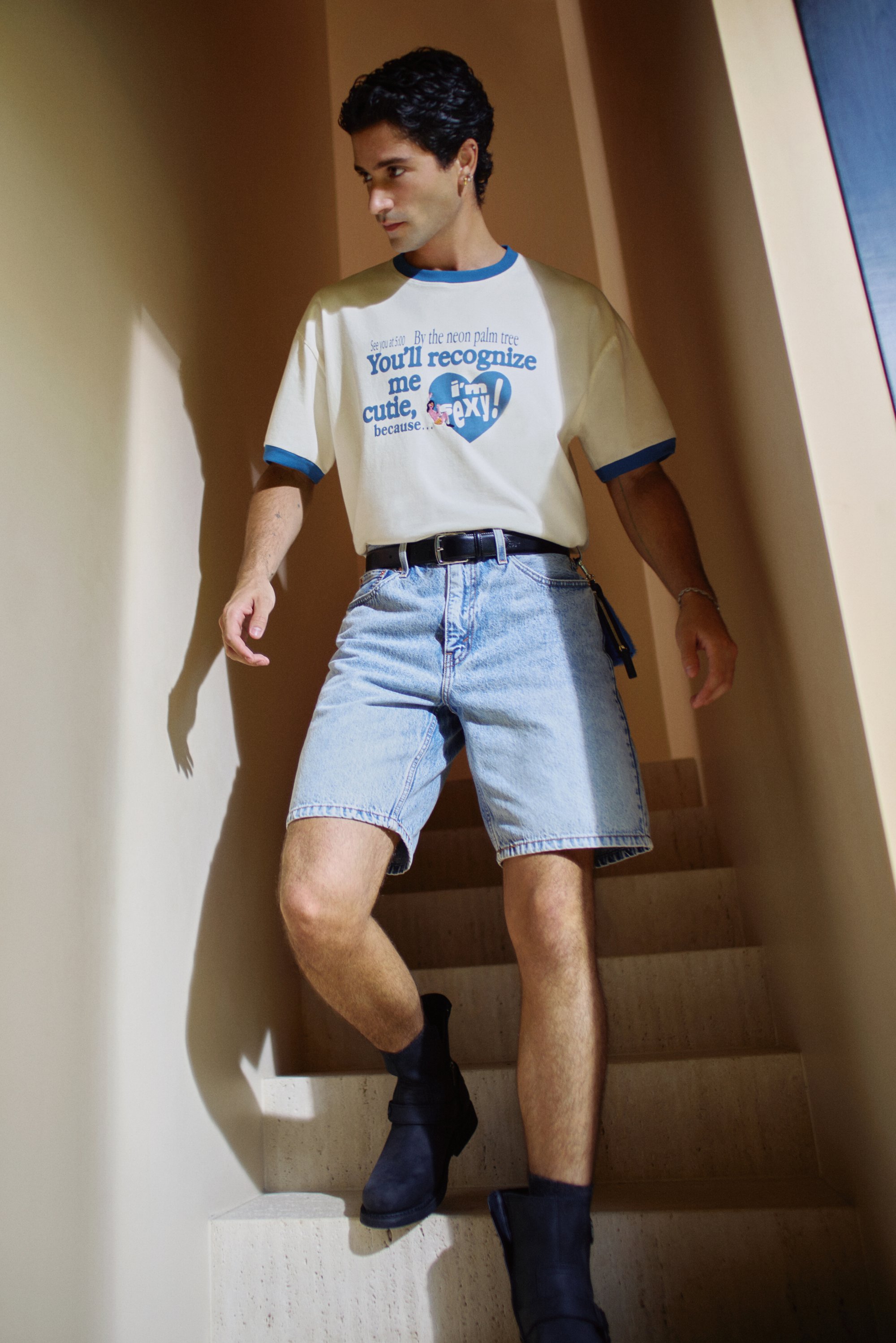 Lässiges Outfit mit einem cremefarbenen T-Shirt mit blauen Akzenten und grafischem Text, kombiniert mit hellblauen Denim-Shorts und schwarzen Ankle Boots.