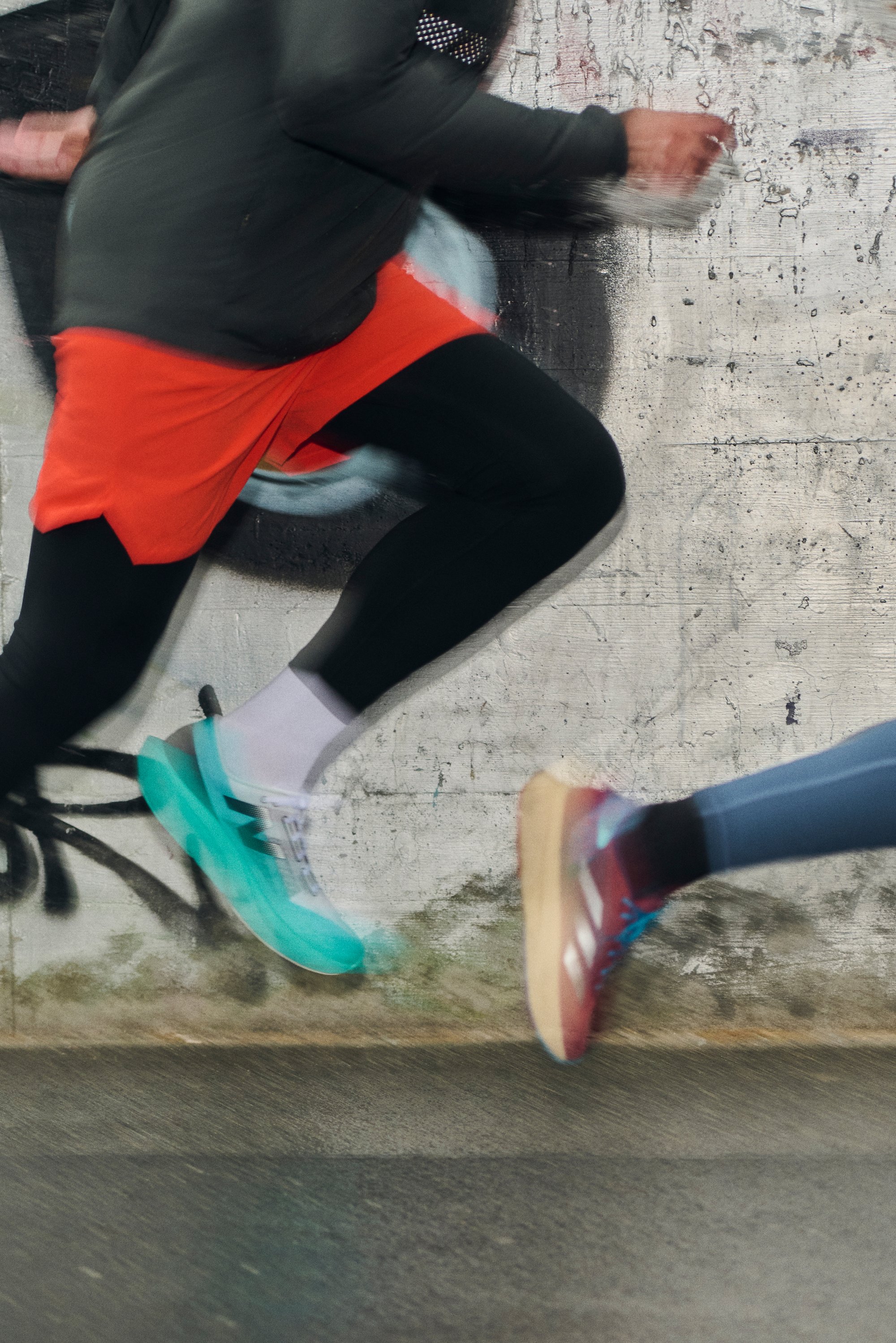 Two people in motion jump on a pavement, wearing colorful athletic gear and shoes. A graffiti-covered wall is visible in the background.