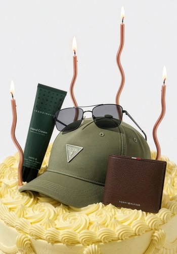 Gâteau jaune givré surmonté d'une casquette Guess verte, de lunettes de soleil noires, d'un portefeuille Tommy Hilfiger marron, d'une crème pour les mains Skandinavisk verte, et de quatre bougies torsadées allumées.
