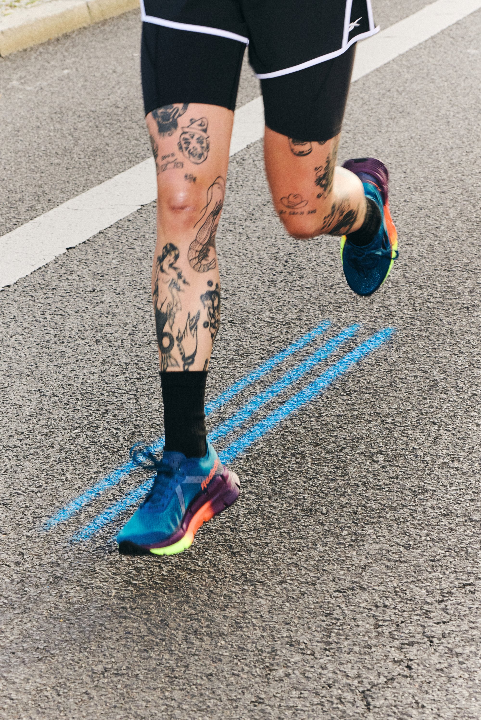 A runner in black shorts and colorful shoes jogs on a paved road, showcasing heavily tattooed legs and blue painted lines beside them.