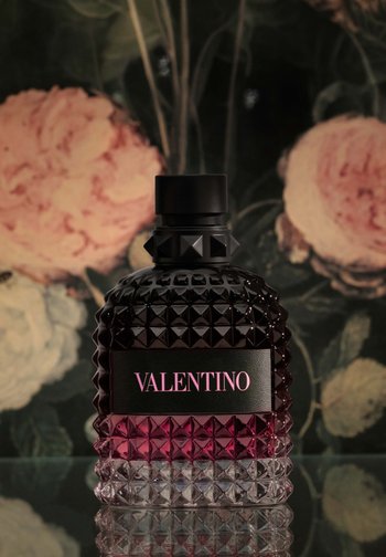 Botella de perfume Valentino negra y rosa con tachuelas en forma de pirámide texturizadas, colocada sobre una superficie reflectante contra un fondo floral con flores en tonos suaves rosa y crema.