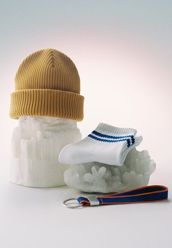 Rumena pletena kapa na belem kristalnem stojalu, bele nogavice z modrimi črtami na manjšem kristalu in modro-oranžen obesek za ključe na beli površini.