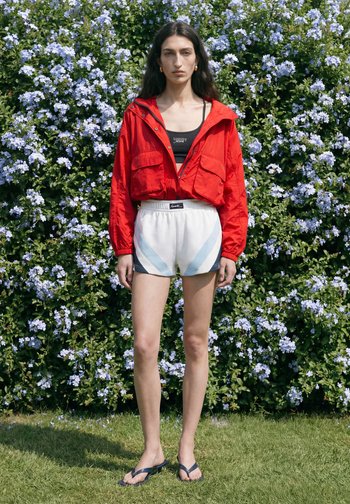 Vrouw staat buiten voor bloeiende struiken, draagt een rode jas, een zwart cropped top, witte korte broek en zwarte slippers.