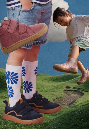 Bambino con scarpe da ginnastica nere con bordo arancione e calzini bianchi con fiori blu, un altro bambino sorride e salta con stivali rosa sull'erba.