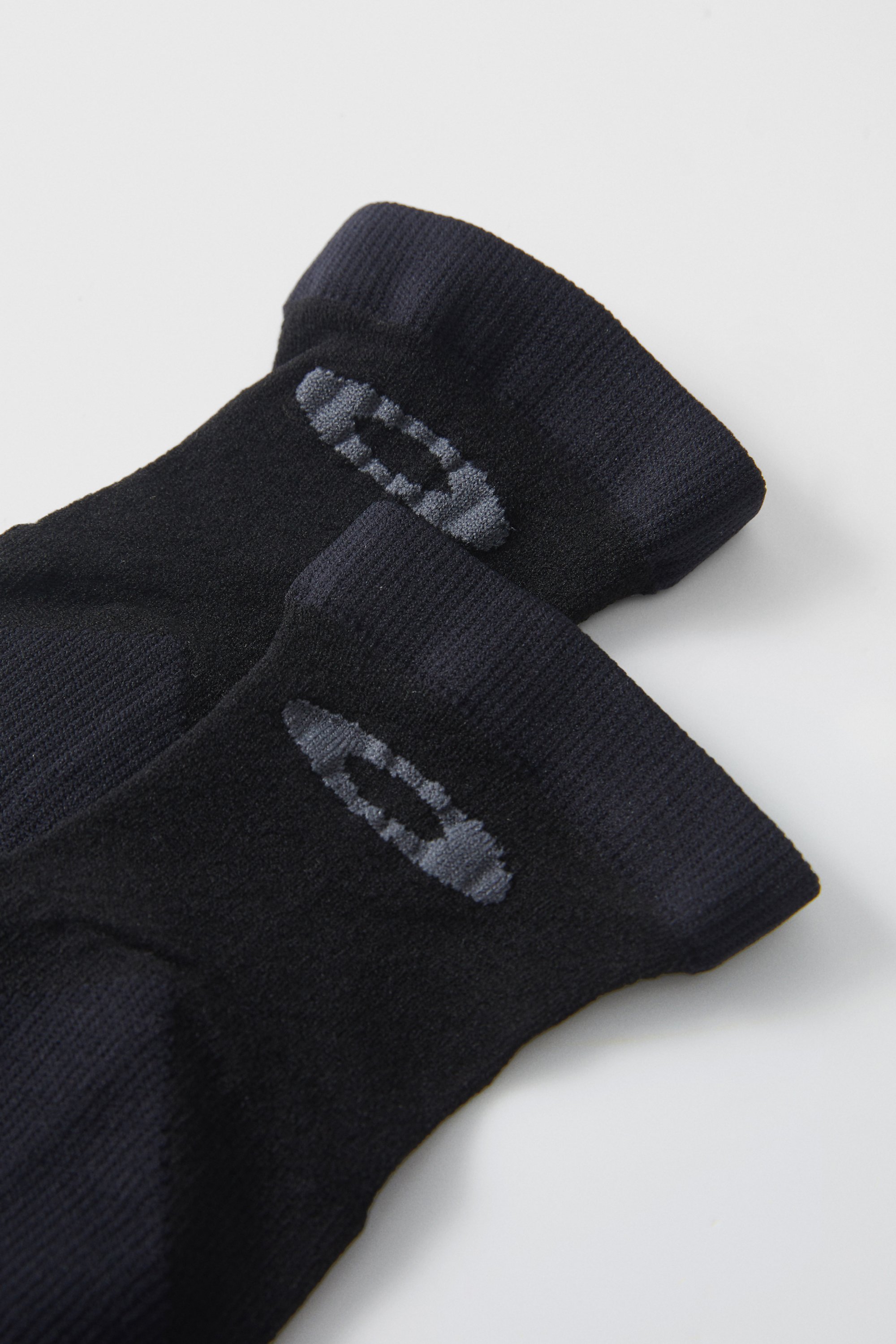 Calcetines negros con logotipos gris claro aparecen uno al lado del otro sobre un fondo blanco, destacando su diseño y contraste de color.
