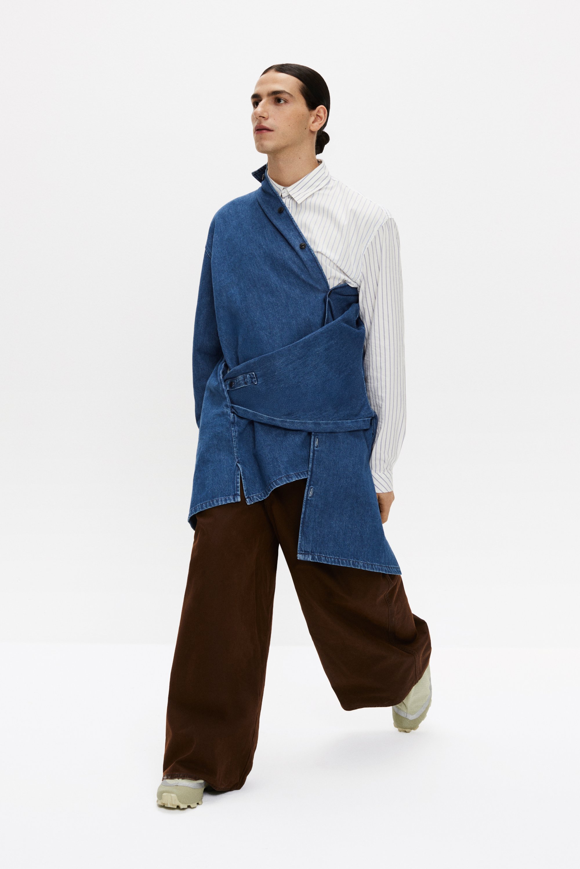 Una modella cammina con sicurezza, indossando un outfit a strati caratterizzato da un top in denim sopra una camicia a righe e pantaloni brown a gamba larga con sneakers.