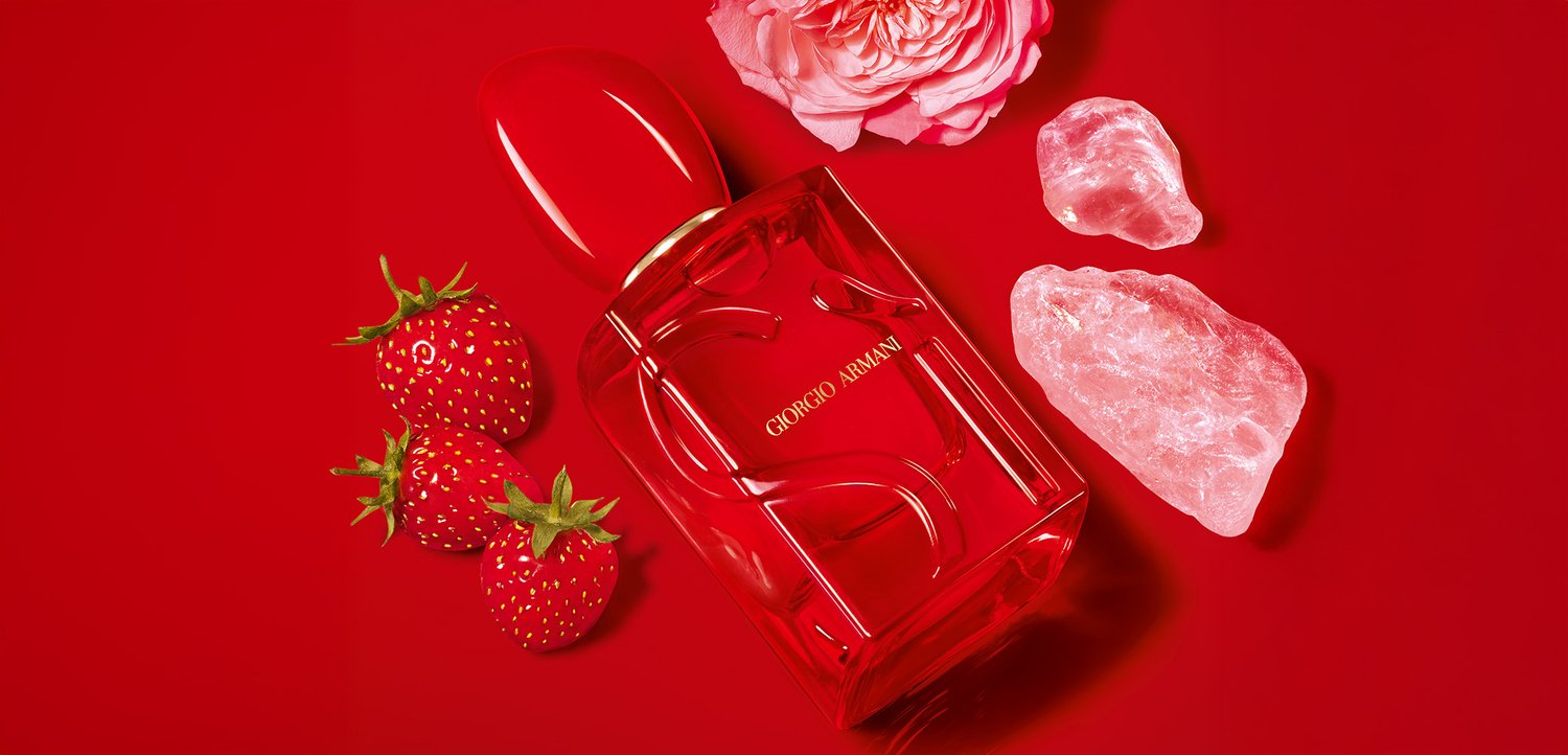Red perfume bottle with a rounded cap, gold lettering, accompanied by strawberries, a pink rose, and crystal-like stones on a red background.