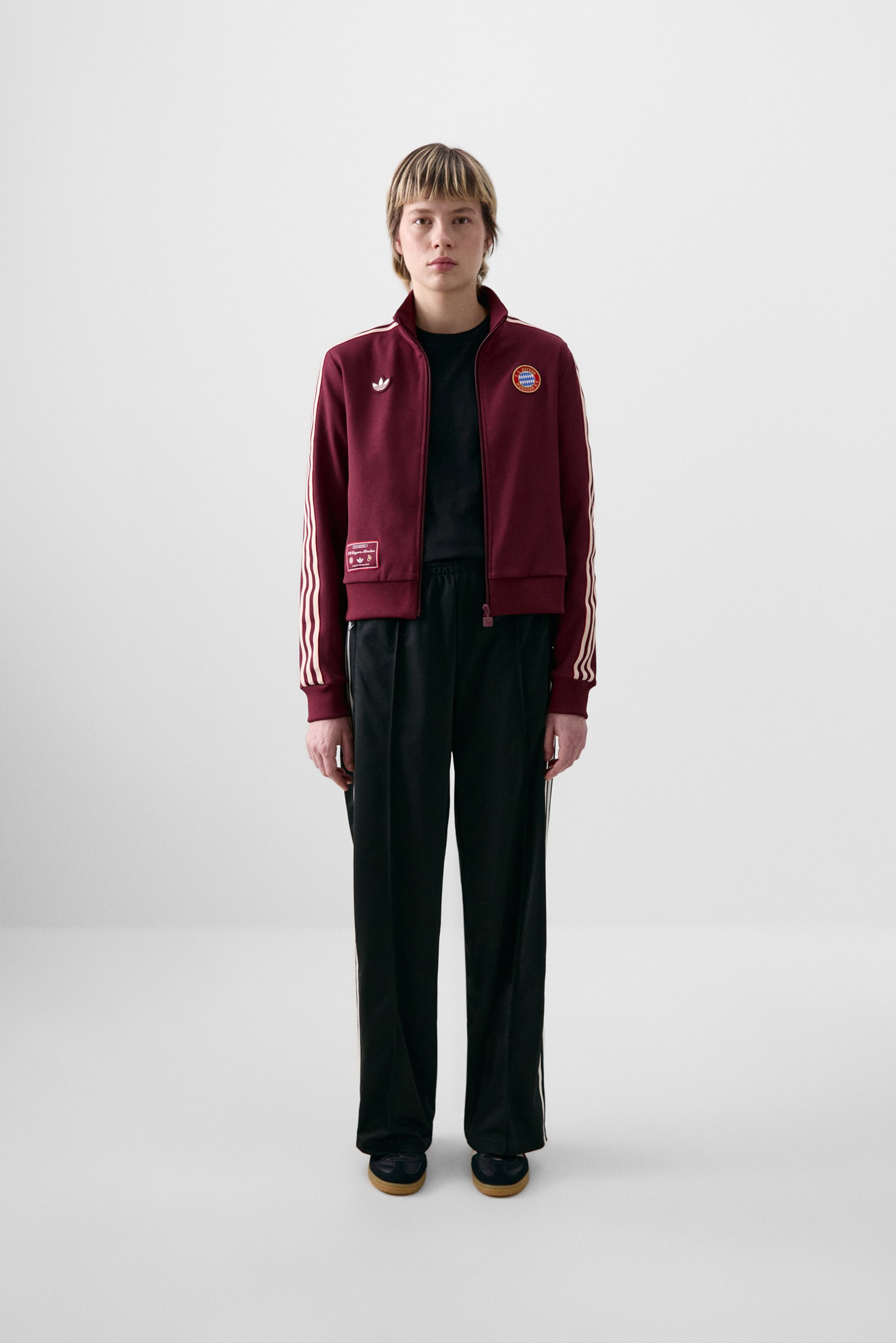 Een persoon staat tegen een effen witte achtergrond, gekleed in een kastanjebruine Adidas zip-up jack, een zwart shirt en zwarte broek.