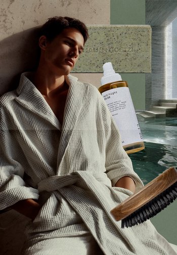 Vit texturerad badrock med knytning i midjan, en klar oljeflaska med vit etikett, en grön tvåltvål och en träborste med svarta borst.