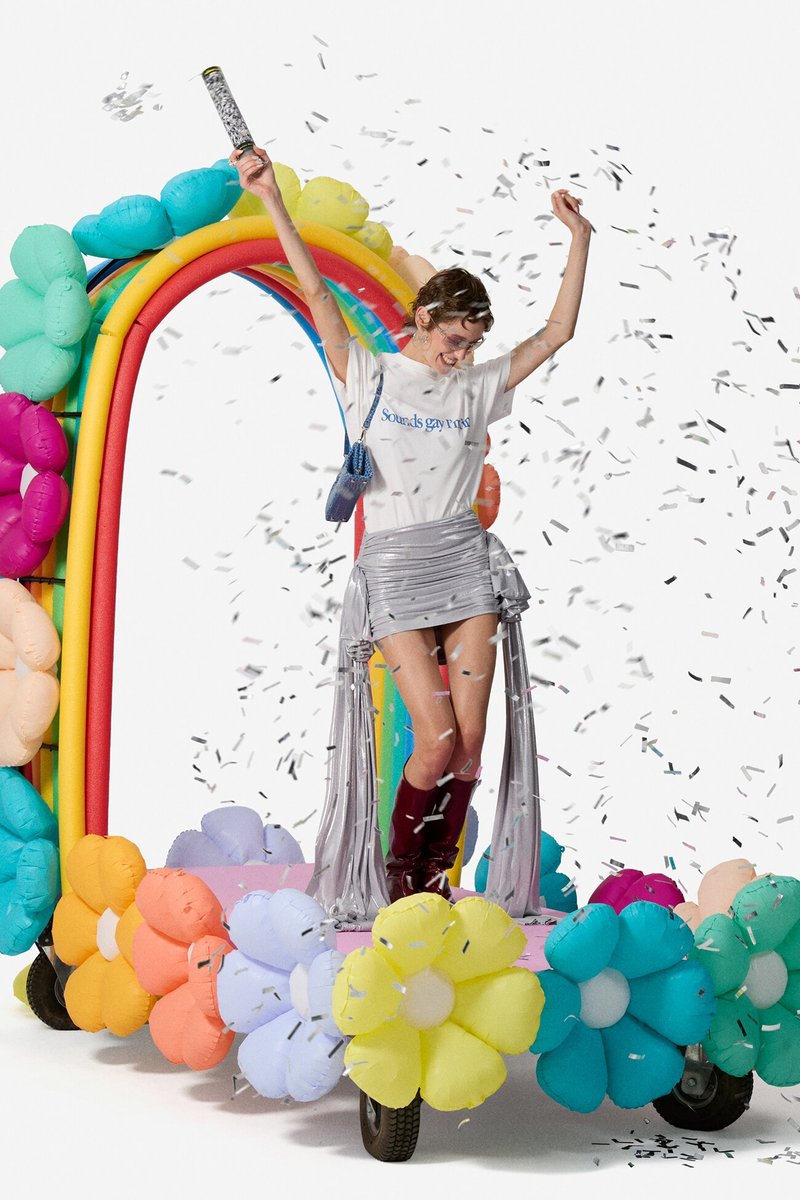 Person celebrating on a colorful flower and rainbow decorated cart, with confetti falling around them and arms raised in joy.