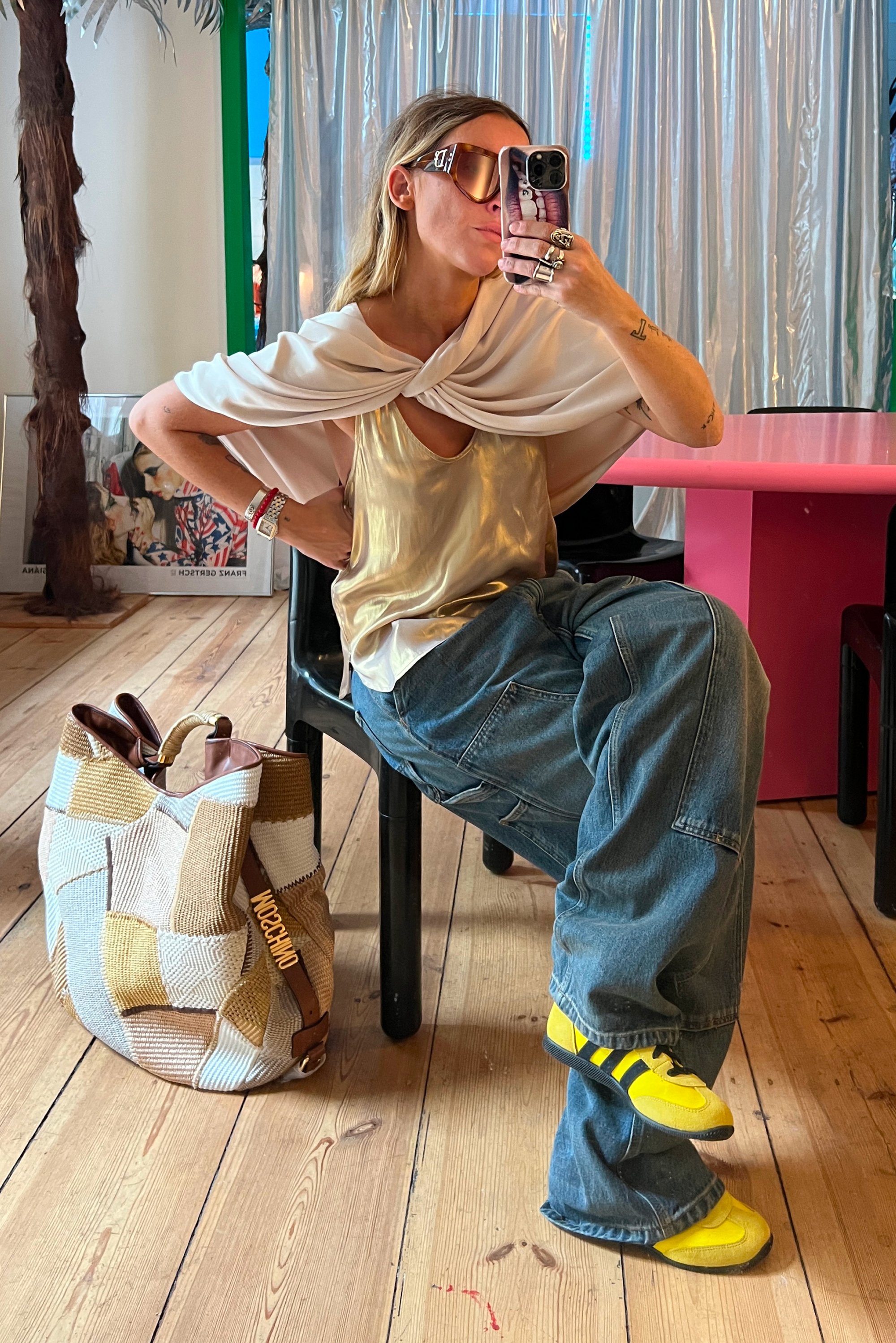Mujer sentada en una silla negra en interiores, vistiendo una camiseta dorada sin mangas, un chal ligero, jeans holgados, zapatillas amarillas, gafas de sol grandes, sosteniendo un smartphone para hacerse un autorretrato en el espejo.