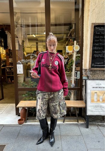 Mujer con suéter rojo de Adidas, pantalones cortos camuflados, botas negras puntiagudas y collares dorados mirando el teléfono fuera de una heladería.