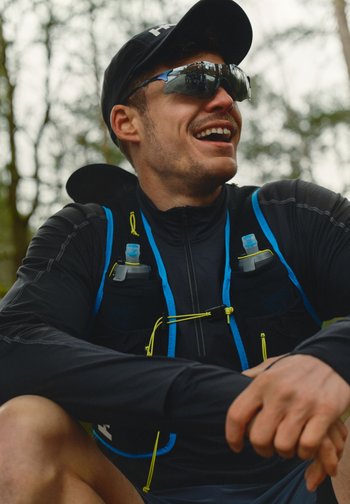 Løper iført svart caps, solbriller og hydreringvest med vannflasker, sittende utendørs og smilende i en skog.