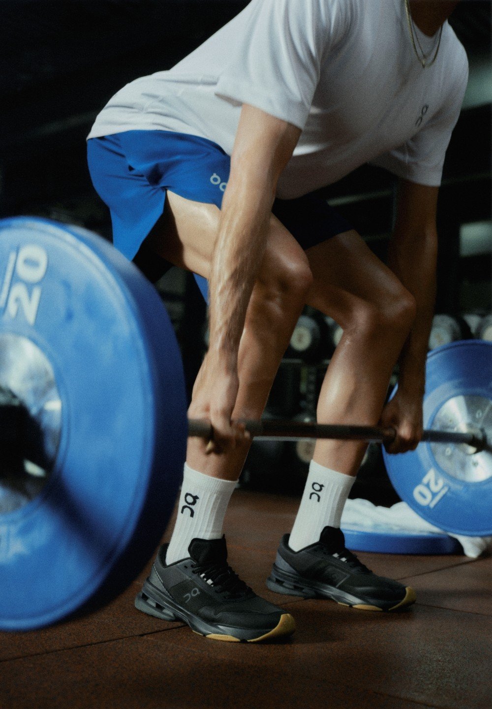 Chaussures de sport noires avec une semelle texturée, chaussettes blanches aux chevilles, shorts bleus et t-shirt gris, en train de soulever une barre avec des disques de poids bleus.