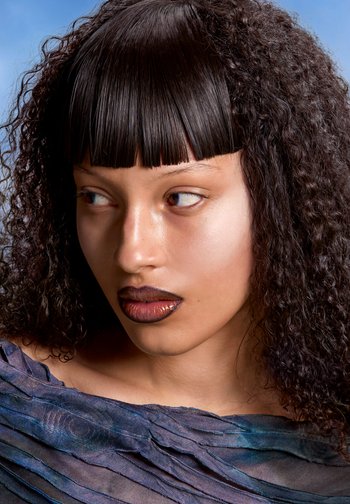 Model with textured black hair and blunt bangs gazes off-camera, wearing dark glossy lipstick and a textured blue fabric top against a pale background.
