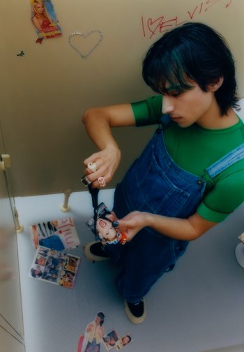 Una persona con una camisa verde y un overol de mezclilla recorta una revista con tijeras, rodeada de imágenes y carteles esparcidos por el suelo.