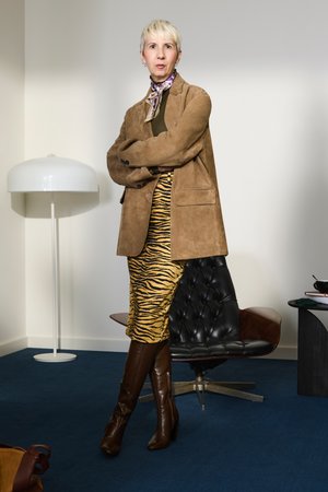 Brauner Wildleder-Blazer über einem grünen Top, kombiniert mit einem gelb-schwarzen Tigerstreifenrock und braunen Kniehosenstiefeln, in einem modernen Raum.