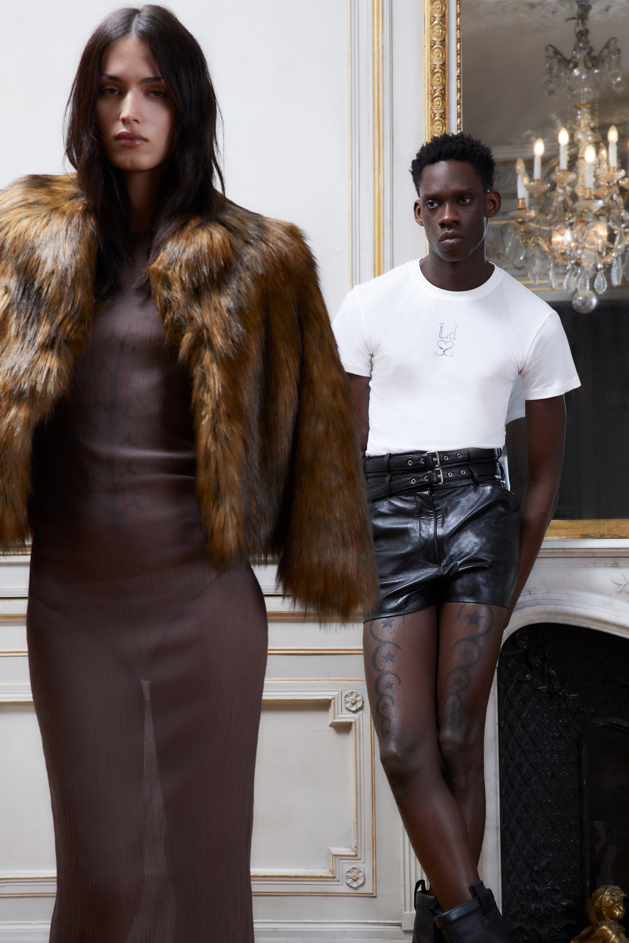 Una modella in un vestito marrone con un cappotto di pelliccia si trova accanto a un uomo in una maglietta bianca e pantaloni di pelle, mostrando la moda in un interno elegante.