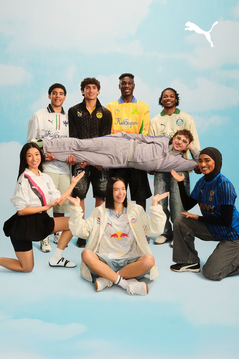 A diverse group of eight young people poses in a playful arrangement, with one person held horizontally above the others against a blue sky backdrop.