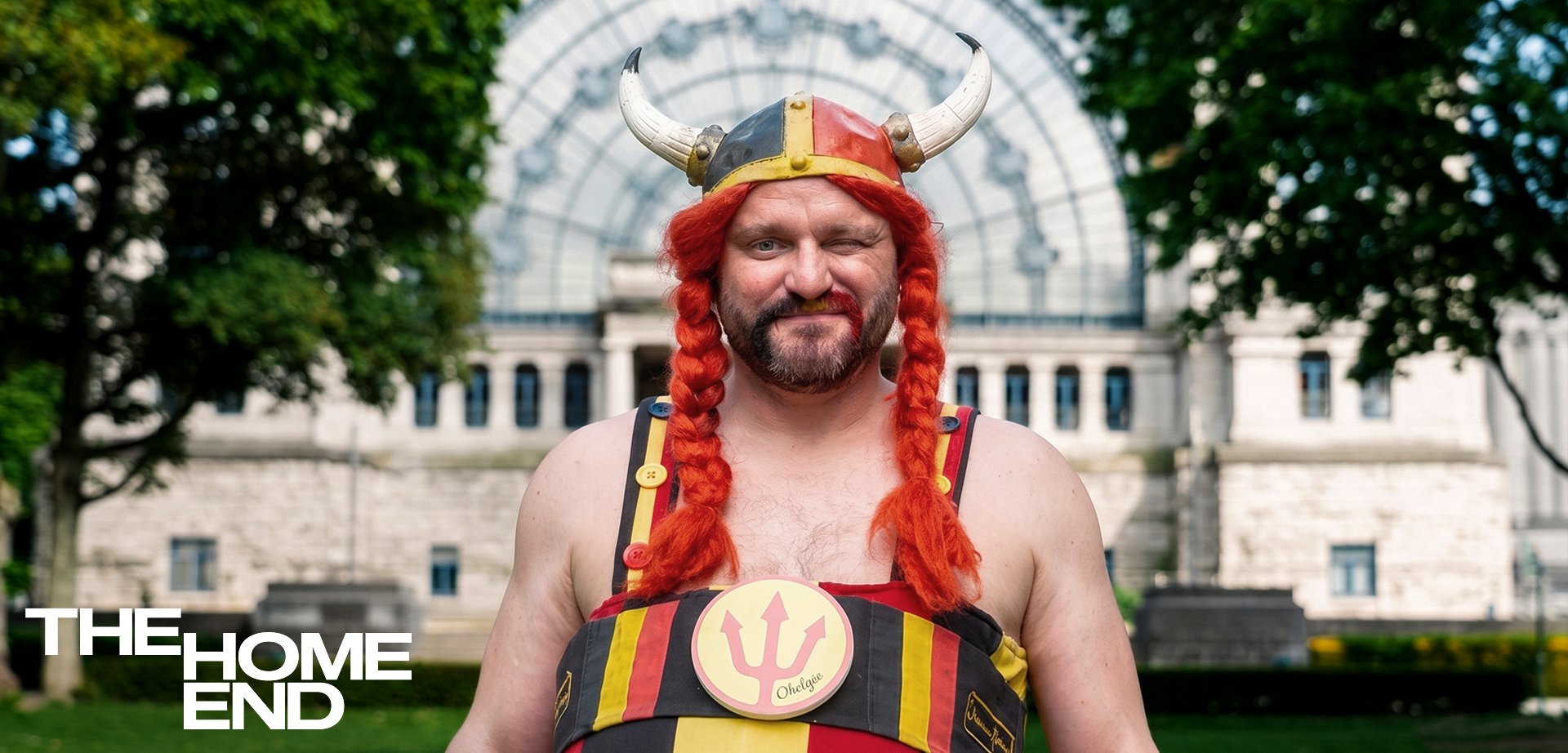 Un homme avec un casque viking, des tresses rouges et une tenue rayée se tient dehors près d'un bâtiment classique, souriant avec un œil fermé.