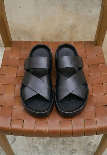 Sandales en cuir noir avec larges brides croisées et semelles épaisses posées sur un siège de chaise en cuir marron tissé.